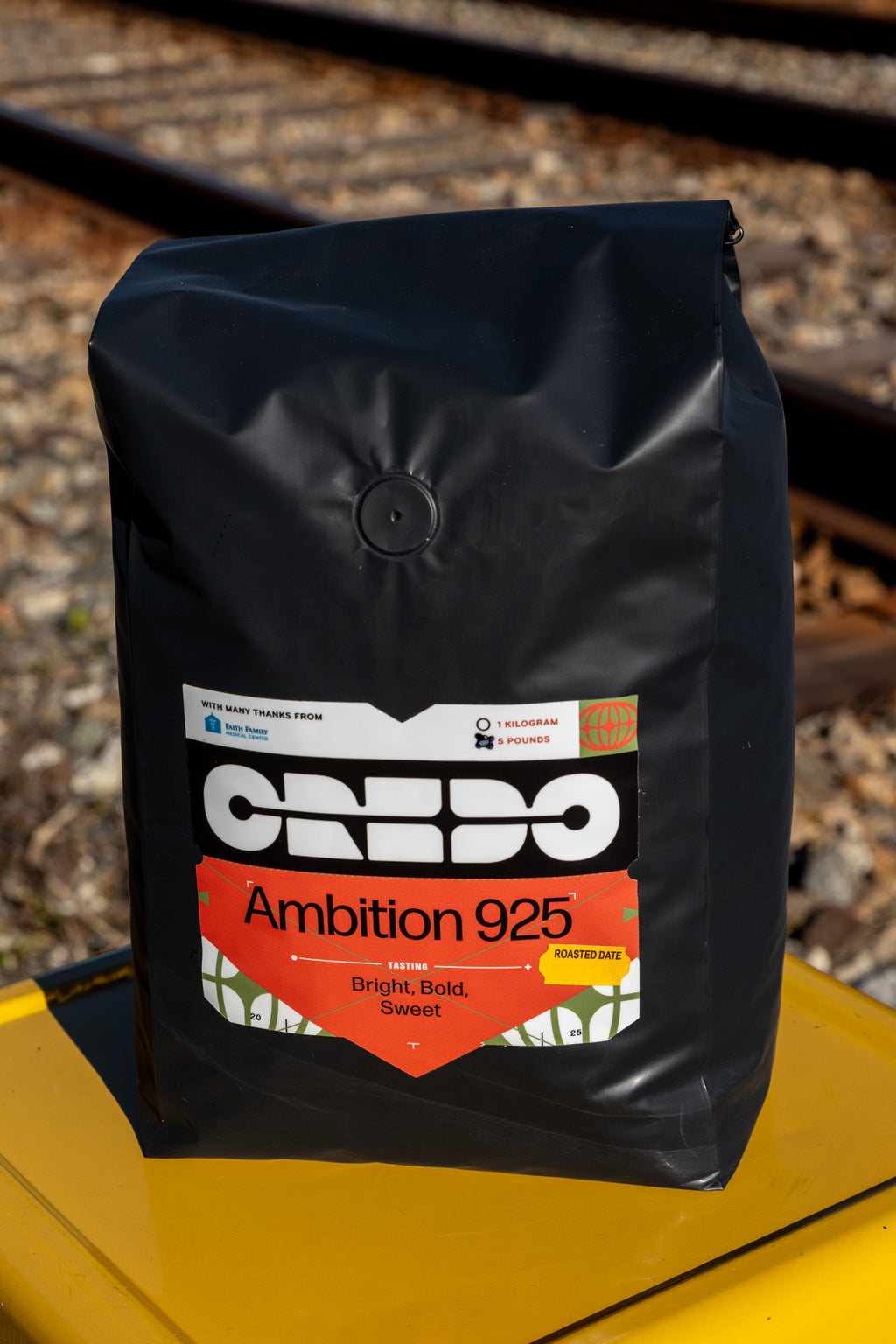 Black coffee bag with 'CREDO Ambition 925' label on a yellow surface with train tracks in the background