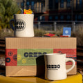 Two white mugs with 'CREDO Coffee Roasters' branding on a cardboard box with the same branding, set against an urban background.