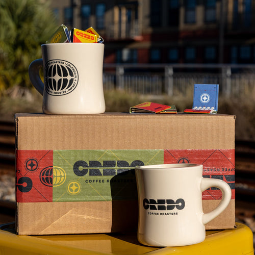 Two white mugs with 'CREDO Coffee Roasters' branding on a cardboard box with the same branding, set against an urban background.