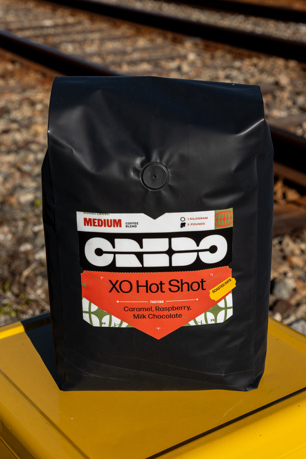 Black coffee bag labeled 'CREDO XO Hot Shot' on a yellow surface with blurred train tracks in the background.